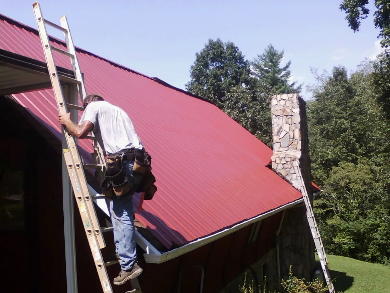Roofer on a ladder beside a red metal roof and stone chimney — Soffit & Fascia Repair by Roofing Brothers in Mount Hood Parkdale